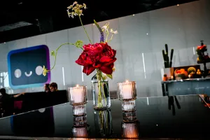 Un arreglo floral de rosa en un jarrón pequeño con agua entre dos velas en una mesa negra alta. Al fondo hay un taco bar a la derecha y un lounge con un gran decal de Houston a la izquierda.