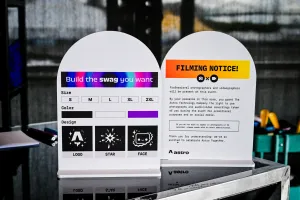 Dos carteles sobre una barra negra. A la izquierda, instrucciones para imprimir tu propia camiseta con tallas, colores, y diseños. A la derecha, un aviso de grabación en inglés.