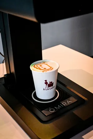 Máquina negra de impresión de arte en café. Debajo de la máquina hay un café con Houston impreso encima.