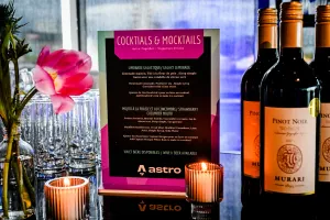 Menú de cocktails y mocktails sobre una barra negra adornada con arreglos florales, velas, y botellas de vino.