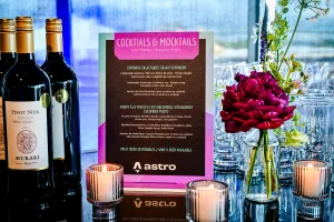 Menú de cocktails y mocktails sobre una barra negra adornada con arreglos florales, velas, y botellas de vino.