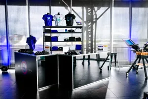 Dos barras negras con apparel mostrado: camisetas púrpura, blancas, y negras así como riñoneras negras de marca. A la derecha, una prensa de impresión de camisetas.