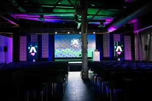 Vista desde el fondo del área oscura y vacía de presentación de la keynote. Filas de sillas negras vacías. Una gran pantalla LED vibrante con el logo de Astro Together. Dos pantallas más pequeñas en orientación retrato adornan los lados del escenario, también con el logo de Astro Together.
