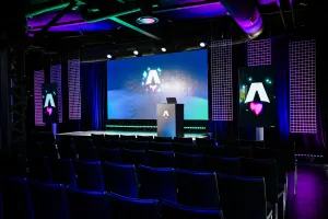 Vista desde el fondo del área oscura y vacía de presentación de la keynote. Filas de sillas negras vacías. Una gran pantalla LED vibrante con el logo de Astro Together. Dos pantallas más pequeñas en orientación retrato adornan los lados del escenario, también con el logo de Astro Together.