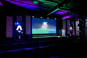 Vista desde el fondo del área oscura y vacía de presentación de la keynote. Filas de sillas negras vacías. Una gran pantalla LED vibrante con el logo de Astro Together. Dos pantallas más pequeñas en orientación retrato adornan los lados del escenario, también con el logo de Astro Together.