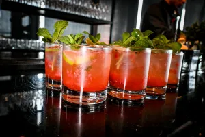 Media docena de cócteles rosas adornados con menta y limón sobre una barra negra.