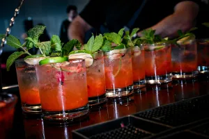 Media docena de cócteles rosas adornados con menta y limón sobre una barra negra.