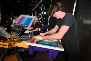 Un hombre usando la prensa de impresión de camisetas para imprimir una camiseta morada para un invitado.