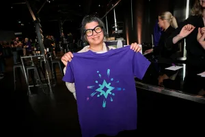 Una mujer con pelo gris y gafas negras sonriendo y sosteniendo una camiseta morada de Starlight.