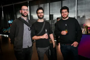 Tres hombres con ropa oscura y gafas posan juntos con bebidas.