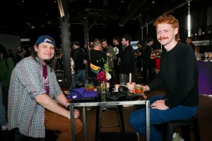 Dos invitados masculinos posan sentados en una mesa alta con su comida, bebidas, y apparel.