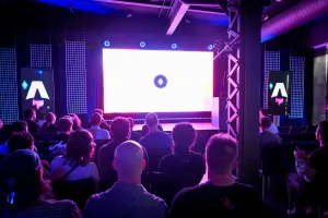 Vista desde el fondo de invitados en sus asientos, esperando a que comience la presentación de la keynote. Están iluminados con luz morada por la gran pantalla LED en el escenario.