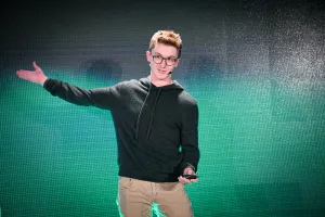Ben Holmes en el escenario frente a una pantalla LED verde y negra. Su brazo derecho está extendido, señalando algo fuera de cámara.