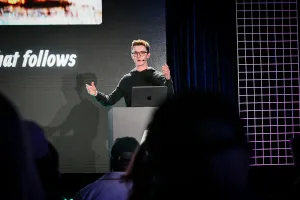 Ben Holmes hablando desde un podio de marca Astro. Tiene ambas manos levantadas mientras se dirige al público.