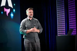 Matthew Phillips hablando en el escenario junto a un podio con un portátil encima. Detrás de él está el logo de Astro Together en pantalla.
