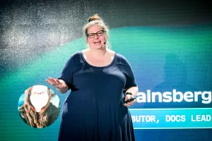 Sarah Rainsberger en el escenario, presentándose. Su mano derecha gesticula hacia el público mientras la izquierda sostiene un clicker.