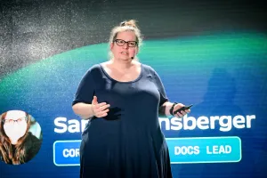 Sarah Rainsberger en el escenario, presentándose. Su mano derecha gesticula hacia el público mientras la izquierda sostiene un clicker.