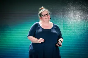 Sarah Rainsberger hablando en el escenario frente a una pantalla LED verde y negra.