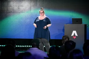 Sarah Rainsberger hablando en el escenario junto a un podio de marca Astro, frente a una pantalla LED verde y negra.