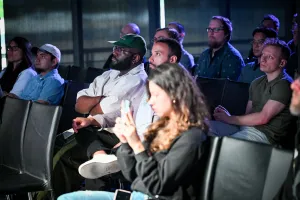Miembros del público escuchando a Fred K. Schott hablar en el escenario. Una mujer levanta su teléfono, grabando contenido de social media de la keynote.