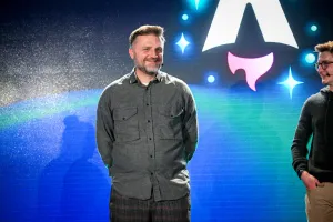 Primer plano de Matthew Phillips de pie con otros oradores de la keynote en el escenario. Tiene las manos detrás de la espalda y está sonriendo al público. A la derecha, Ben Holmes le está sonriendo.