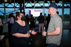 Sarah Rainsberger, llevando una máscara negra, charla con un invitado. El stand de Sentry y una pantalla de TV se pueden ver detrás de ellos.