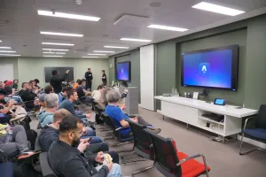 Una gran sala de reuniones con el logo de Astro mostrándose en los monitores con decenas de personas en sillas, esperando que comience la presentación.
